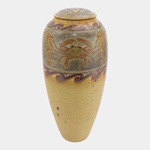 May include: A tall, tapered ceramic urn with a lid, featuring a speckled tan base and a grey upper section decorated with embossed seahorses and a central crab. The design includes wavy purple bands and a circular lid with a detailed motif.