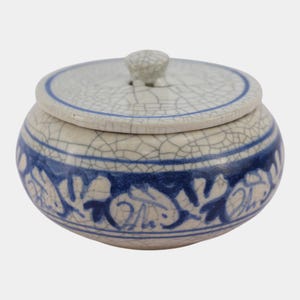 May include: A small, round ceramic jar with a lid. The jar is off-white with a blue floral design around the base and a blue band near the top. The lid has a small knob and a crackle glaze finish.