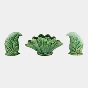 May include: A ceramic centerpiece set featuring a large, scalloped bowl and two leaf-shaped candle holders. The set is glazed in a glossy green with lighter green accents, creating a nature-inspired aesthetic. The bowl is approximately 8 inches in diameter.