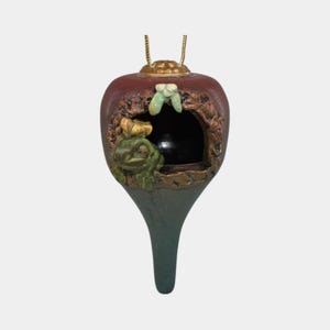 May include: A decorative ornament shaped like a bulbous fruit, featuring a carved frog inside a hollowed-out section. The ornament has a dark red top, a green-gray base, and gold accents. A small, green, leaf-like detail is above the frog.