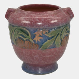 May include: A handcrafted, ceramic vase with a speckled, reddish-pink exterior. The vase features a band of blue with green leaves and orange floral designs. It has two small handles and a blue base.