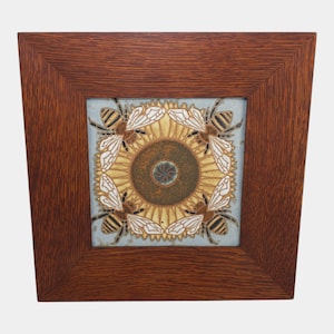 May include: A square ceramic tile framed in dark brown wood. The tile features a central circular motif with a dark textured center and a yellow sunflower-like pattern. Four bees with white wings and striped bodies are positioned in the corners.