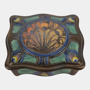 May include: A square ceramic box with a wavy edge, featuring a central circular medallion with orange and brown feather-like patterns. The surrounding surface is decorated with green, blue, and yellow peacock-inspired motifs on a dark brown base.
