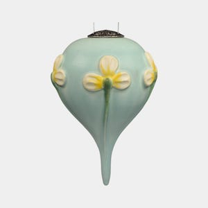 May include: A light blue, teardrop-shaped ornament with three white and yellow flower designs. The ornament has a silver-colored cap and a small loop for hanging. The overall design is elegant and floral.