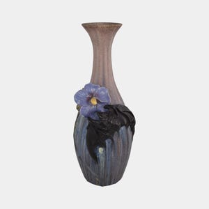 May include: A tall, decorative vase with a slender neck and a wide base. The vase is primarily a muted purple color, with a blue flower and black leaves adorning the front. The flower has a yellow center.