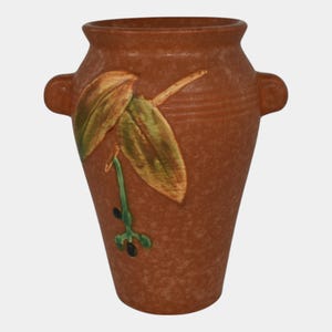 May include: A brown ceramic vase with two side handles and a textured surface. The vase features a painted design of green and yellow leaves with black accents. The vase is a decorative item.