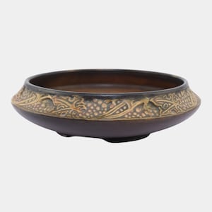 May include: A brown ceramic bowl with a decorative rim featuring a raised pattern of vines and grapes. The bowl has a wide, shallow shape and a dark interior. The rim has a gold and brown color scheme.