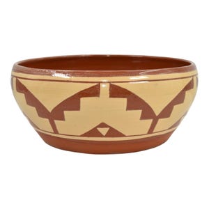 May include: A round, terracotta-colored bowl with a cream-colored interior and geometric, stepped patterns in a darker brown. The bowl has a wide rim and a simple, earthy design.