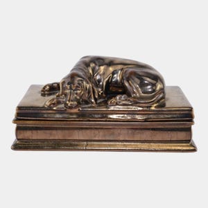 May include: A bronze-colored decorative box with a sculpted dog resting on top. The dog is lying down, with its head and paws resting on the box. The box has a rectangular shape with a textured surface.