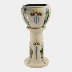 May include: A cream-colored ceramic plant stand with a pedestal base. The stand features colorful geometric designs in blue, green, yellow, and brown. The top of the planter has a dark green interior.