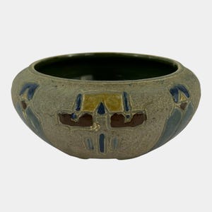 May include: A small, round ceramic bowl with a textured, light gray exterior. The bowl features a dark green interior and is decorated with geometric patterns in shades of blue, brown, and yellow. The bowl is approximately 6 inches in diameter.