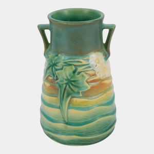 May include: A ceramic vase featuring a two-tone green and yellow glaze with a relief pattern of large leaves and white flowers. The vessel has a tapered body, a wide circular opening, and two small handles extending from the upper neck area.