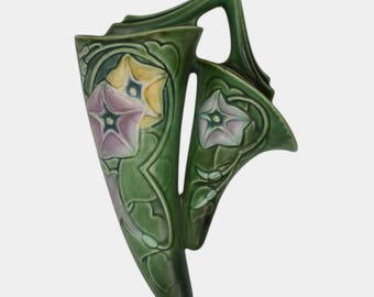 May include: Green ceramic wall pocket vase with a floral design. The vase features stylized flowers in yellow, pink, and white, with green leaves and stems. It has a handle and a unique shape. The vase is likely for decorative purposes.