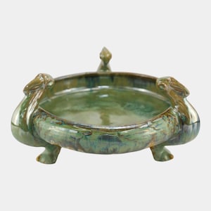 May include: A green ceramic bowl with a glossy finish. The bowl has a rounded shape with three feet and decorative elements on the sides. The bowl is approximately 10 inches in diameter.