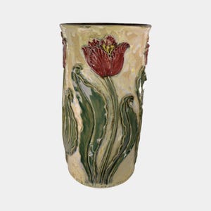 May include: A tall, ceramic vase with a tulip design. The vase features a cream-colored background with a large red tulip and green leaves. The vase has a textured surface and a dark rim.