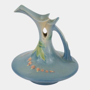 May include: A ceramic pitcher featuring a wide, flat base and a tall, slender neck with a prominent, elongated spout. The surface is finished in a matte blue glaze with soft green leaf accents and a delicate trail of small, orange-colored berries.