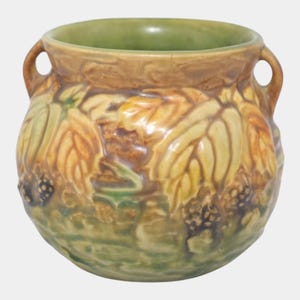 May include: A ceramic vase with a round shape and two handles. The vase features a relief design of leaves and berries in shades of yellow, brown, and green. The interior rim is a light green color.