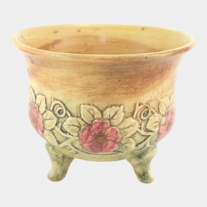 May include: A ceramic planter with a light brown and yellow exterior. The planter features a floral design with pink flowers and green leaves. It has three small feet and a wide rim.