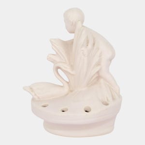 May include: A cream-colored ceramic figurine depicting a child figure with a swan. The child is positioned near the swan, which is surrounded by stylized foliage. The base has three holes, suggesting a possible function as a candle holder or incense burner.