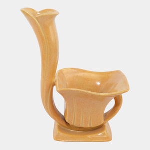May include: A tan ceramic vase with a unique, curved design. The vase features a tall, slender, curved neck that extends from a square base. The main body has a square bowl shape with a handle. The vase is a solid tan color.
