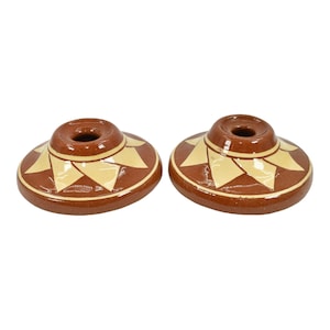 May include: Two terracotta candle holders with a geometric design in a light yellow color. The holders are round and have a small hole in the center for a candle.