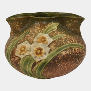 May include: A ceramic vase with a speckled brown and green exterior. The vase features a design of white and yellow daffodils with green leaves. The rim of the vase has a wavy edge.