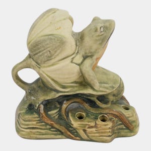 May include: A ceramic frog figurine perched on a lily pad atop a textured base with two circular openings. The piece features muted green, brown, and cream tones with a glazed finish, depicting a frog with its head tilted upward and a handle.