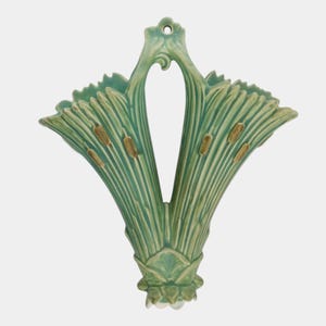 May include: A light green ceramic wall pocket vase featuring a dual-flared design with molded cattail plant details. The piece has a central opening at the top for hanging and a textured, ribbed surface finish throughout the entire structure.
