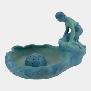May include: A matte blue and teal ceramic flower frog bowl featuring a sculpted figure kneeling on a rock formation at the edge. The bowl has a wavy, organic rim and a central dome-shaped flower frog with multiple holes for arranging stems.