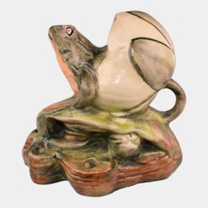 May include: A ceramic frog-shaped teapot with a light green and brown color scheme. The frog is perched on a leaf-shaped base, with the teapot forming the frog's body. The teapot has a handle and a spout.