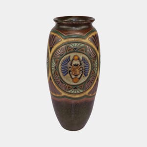 May include: A tall, decorative ceramic vase with a dark brown base and rim. The vase features a central circular design with a scarab beetle motif in blue, green, and gold. The design is framed by geometric patterns and shell-like shapes.