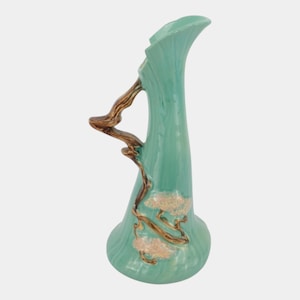 May include: A tall, light turquoise ceramic vase with a flared rim and a glossy finish. The vase features a raised, brown, branch-like handle extending from the side, with textured, light pink floral accents molded onto the lower body of the piece.