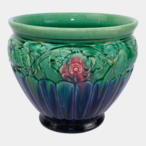 May include: A ceramic planter featuring a glossy finish with a gradient from a deep navy blue base to a vibrant green upper section. The exterior is decorated with a raised relief pattern of flowers and leaves, with pink accents on the petals.