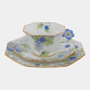 May include: A vintage teacup, saucer, and dessert plate set. The white porcelain set features a floral design with blue flowers and green leaves. The teacup handle is a blue flower. The edges are trimmed with a gold border.