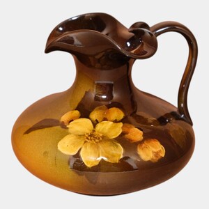 May include: A glossy dark brown ceramic pitcher with a wide, ruffled rim and a curved handle. The body of the pitcher features a hand-painted design of yellow and orange flowers with green leaves, set against a deep brown, shaded background.