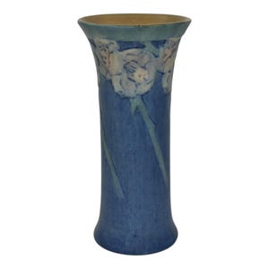 May include: A blue ceramic vase with a flared top and a cylindrical body. The vase features embossed white floral designs around the rim and light blue stems that run down the body. The vase is approximately 10 inches tall.