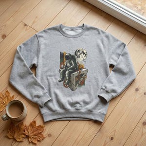 Könnte beinhalten: Ein graues Sweatshirt mit einem grafischen Aufdruck eines Astronauten, der auf einer Struktur mit Zahnrädern, einem Mond und Himmelselementen sitzt. Das Design verwendet Grau-, Braun- und Gelbtöne. Das Sweatshirt hat einen Rundhalsausschnitt und lange Ärmel.