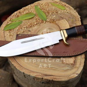 Può includere: Un grande coltello in acciaio lucido con un manico nero e marrone e dettagli dorati, appoggiato su un fodero in pelle marrone. Il coltello è esposto su una superficie di legno con foglie verdi. Il testo "ExpertCraft ART" è visibile.