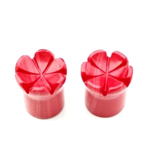 Könnte beinhalten: Zwei rote, blütenförmige Ohrstöpsel. Jeder Stöpsel hat einen zylindrischen Sockel und eine Oberseite, die in ein sechszackiges Blumendesign geschnitzt ist. Die Stöpsel sind einfarbig, glänzend rot mit subtiler Marmorierung.