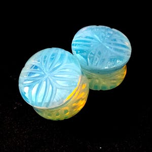 May include: Two light blue, carved gemstone cabochons with a floral design. The gemstones are round and have a reflective surface, with yellow and orange hues visible in the reflection. The background is black.