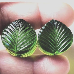 May include: A pair of dark green ear plugs featuring a carved leaf design with prominent veins. The translucent, polished surface of the plugs catches the light, highlighting the detailed, textured ridges that mimic the appearance of a natural leaf.