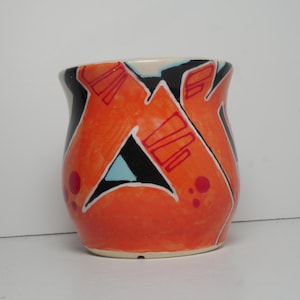 May include: A ceramic mug featuring a vibrant orange base color with bold black, white, and light blue geometric graffiti-style patterns. The design includes sharp angular shapes and small red circular accents across the exterior surface.