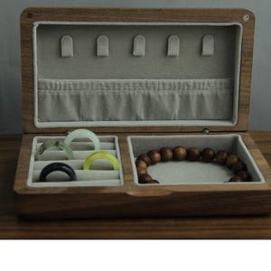 May include: A wooden jewelry box with a light gray interior and multiple compartments. The box contains rings in various colors and a beaded bracelet. The box is open, revealing the contents, and is made of a dark wood.