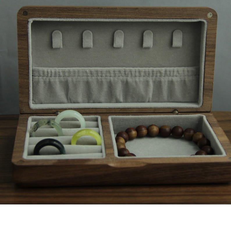 May include: A wooden jewelry box with a light gray interior and multiple compartments. The box contains rings in various colors and a beaded bracelet. The box is open, revealing the contents, and is made of a dark wood.