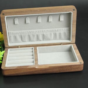 May include: Open wooden jewelry box with a light gray interior. The box has a rectangular shape with a hinged lid. The interior is divided into compartments, including ring slots and earring holders. The box is made of a light brown wood.
