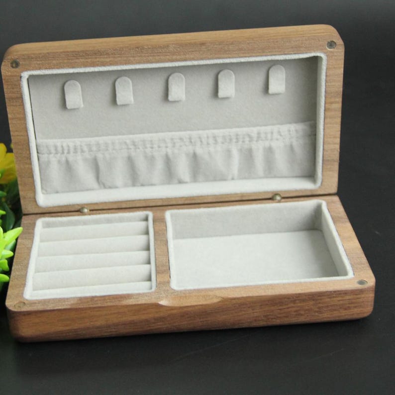 May include: Open wooden jewelry box with a light gray interior. The box has a rectangular shape with a hinged lid. The interior is divided into compartments, including ring slots and earring holders. The box is made of a light brown wood.