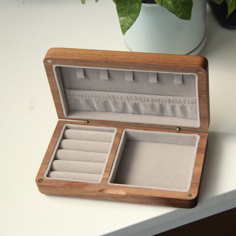 May include: Open wooden jewelry box with a light gray velvet interior. The box is rectangular with a hinged lid and compartments for rings and other jewelry. The wood has a natural grain and a warm brown color.