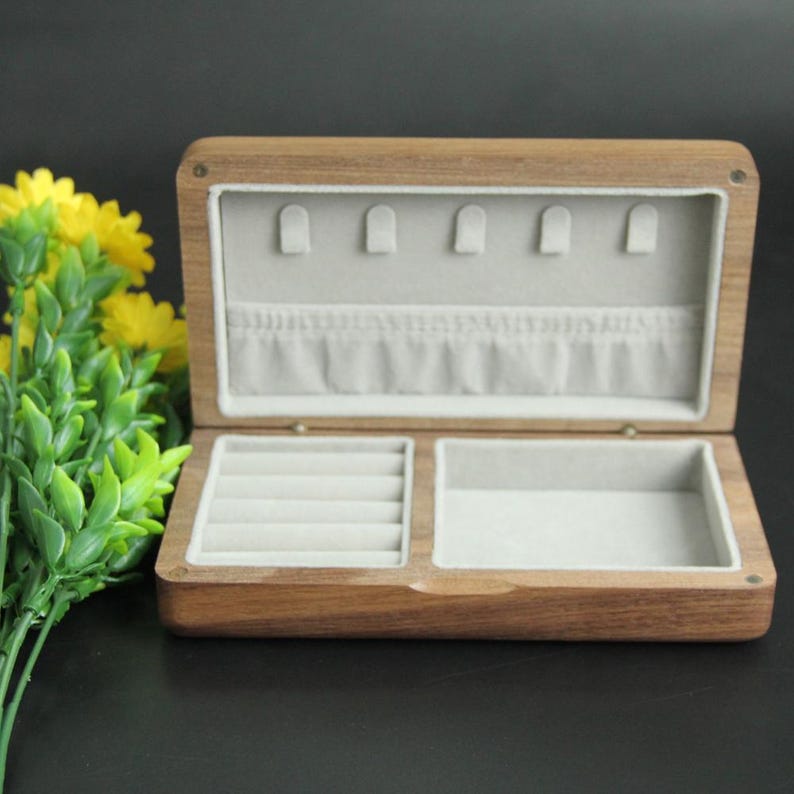 May include: Open wooden jewelry box with a light gray interior. The box is rectangular with a hinged lid and features compartments for rings and other jewelry. The wood has a natural grain and a warm brown tone.