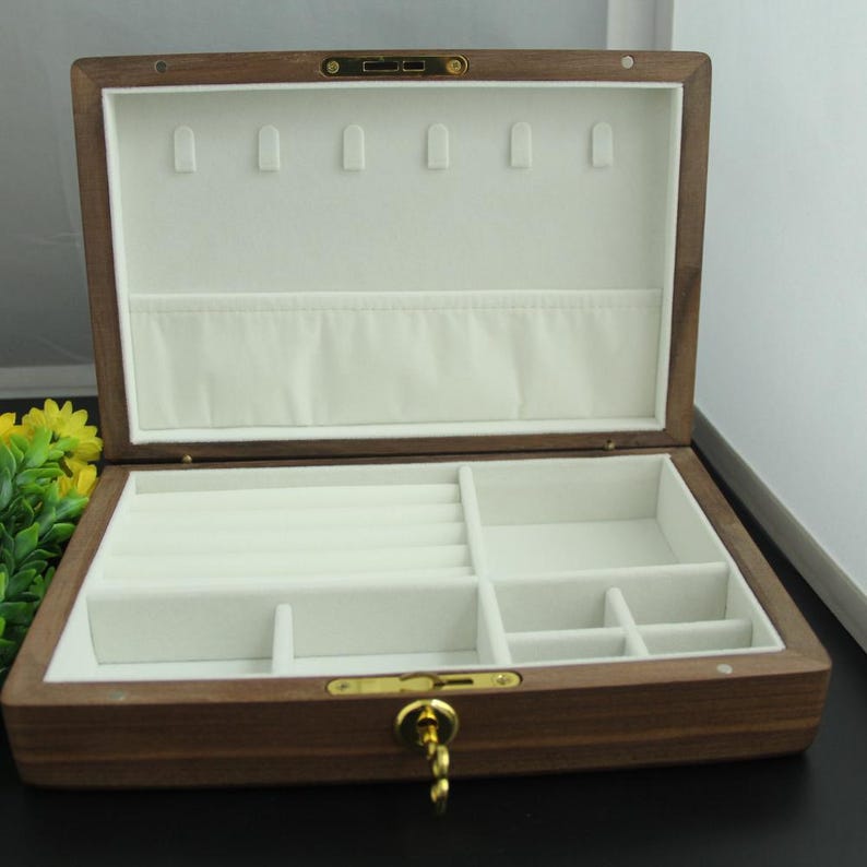 May include: Open wooden jewelry box with a light-colored interior. The box has multiple compartments and slots for organizing jewelry. The exterior is a dark brown color with gold-colored hardware, including a clasp and hinges.