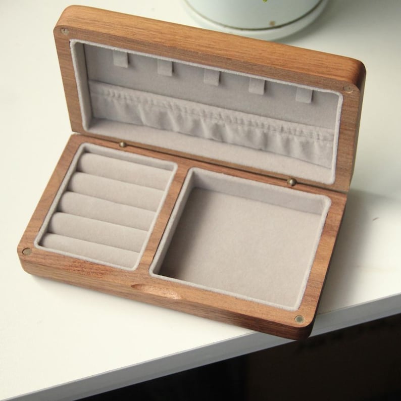 May include: Open wooden jewelry box with a light brown finish. The interior is lined with a light gray fabric, featuring ring slots and a compartment for other jewelry. The box is open, revealing its contents.